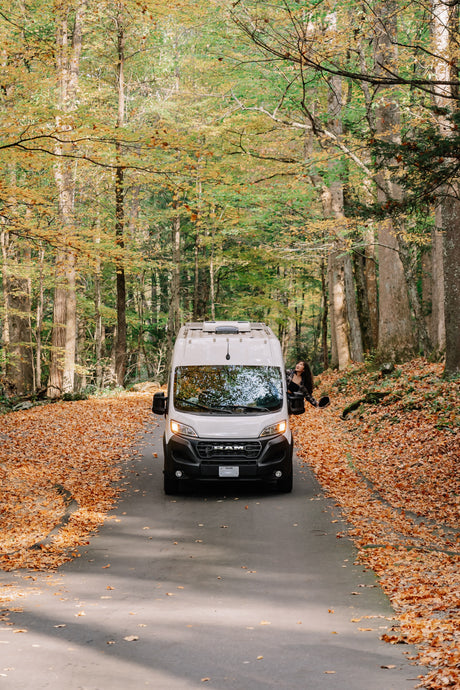 Embrace autumn with an RV rental! A white camper van drives a leaf-covered road in a vibrant fall forest, passenger looking out.