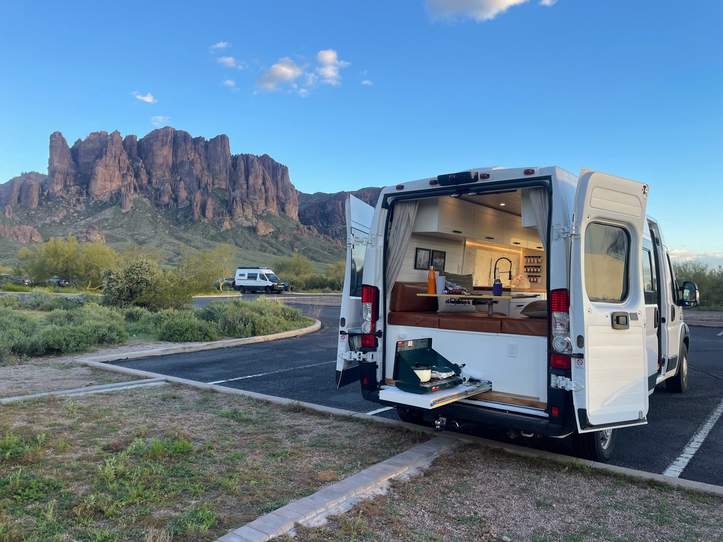 Experience van life! This RV rental features an open campervan with an outdoor kitchen, parked against stunning desert mountains.