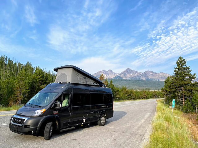 Black RV rental van with pop-top extended, parked roadside. Majestic mountains and forests define this scenic travel.