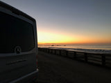 RV rental Airstream van parked on sandy beach at sunset/sunrise. Ocean waves, distant pier, and rocks offer a beautiful travel view.