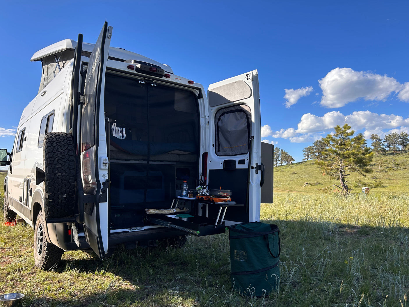 An RV rental with open doors, revealing an organized van life setup for cooking & camping in a scenic grassy field.