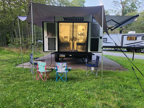 A Winnebago camper with an extended awning and camping chairs is set up at a leafy campsite, ready for an RV rental trip.