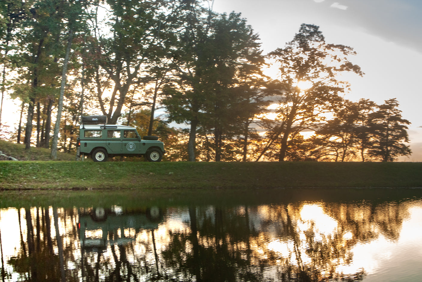 Green Land Rover with rooftop tent beside a lake at sunset. Your ideal **RV rental** for scenic outdoor adventures.