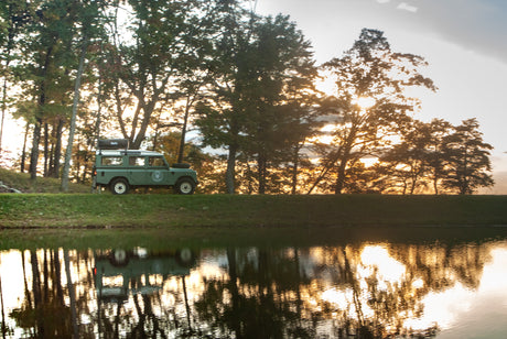 Green Land Rover with rooftop tent beside a lake at sunset. Your ideal **RV rental** for scenic outdoor adventures.
