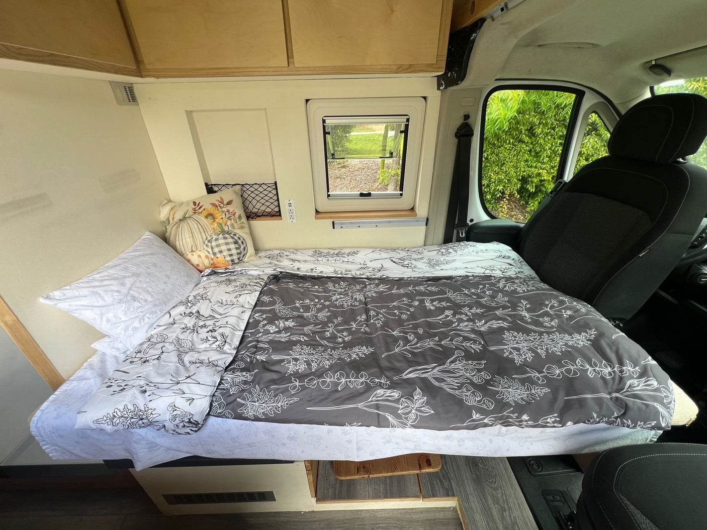 Modern RV rental interior: a cozy bed with decorative pillows, window view, and front cabin with driver's seat.
