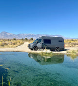 Grey RV rental (Sprinter van) by a clear, turquoise natural spring with mountains. Perfect for scenic nature trips.