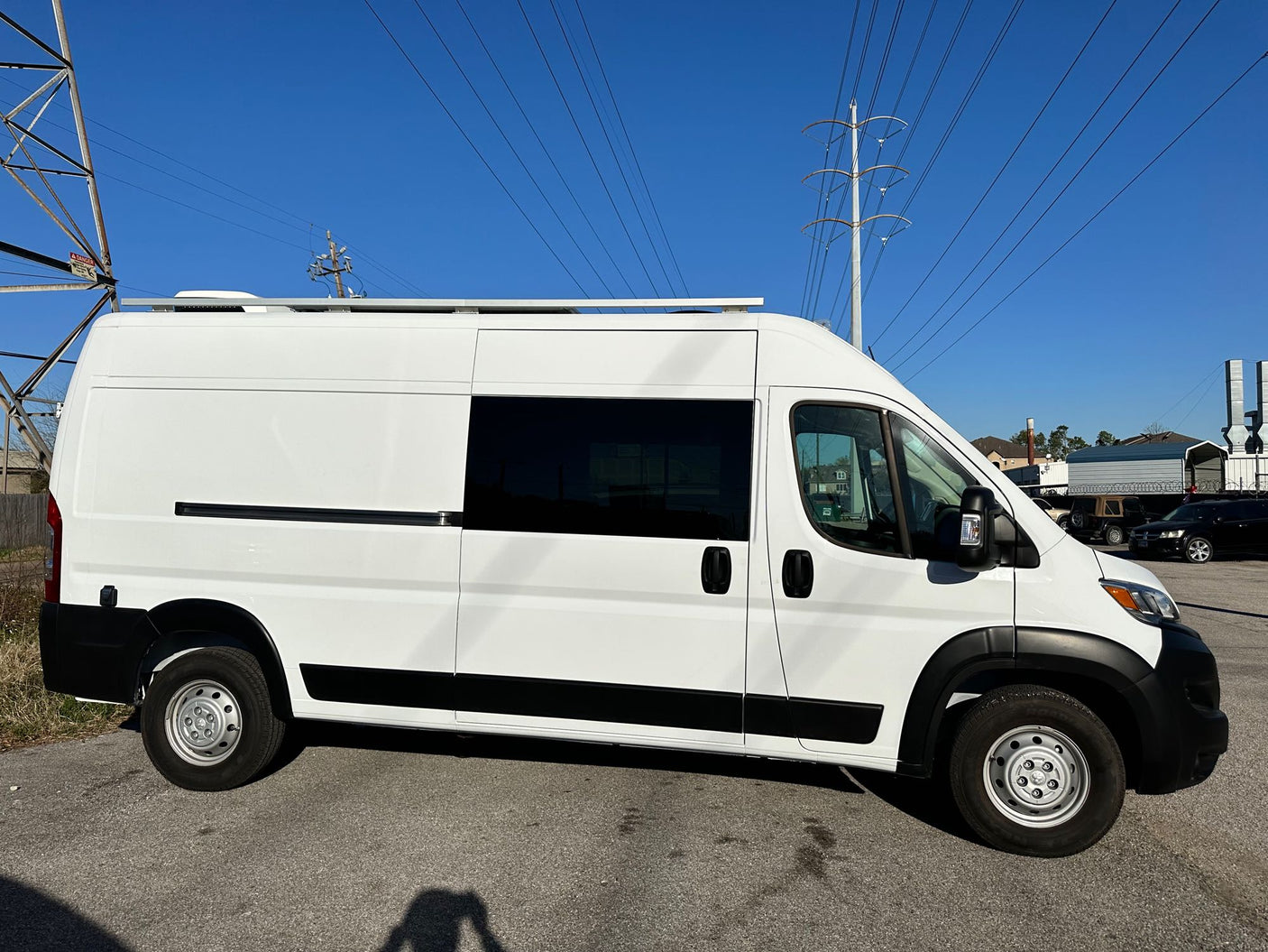 White converted camper van, an RV rental option, with roof rack system parked under power lines on a sunny day.