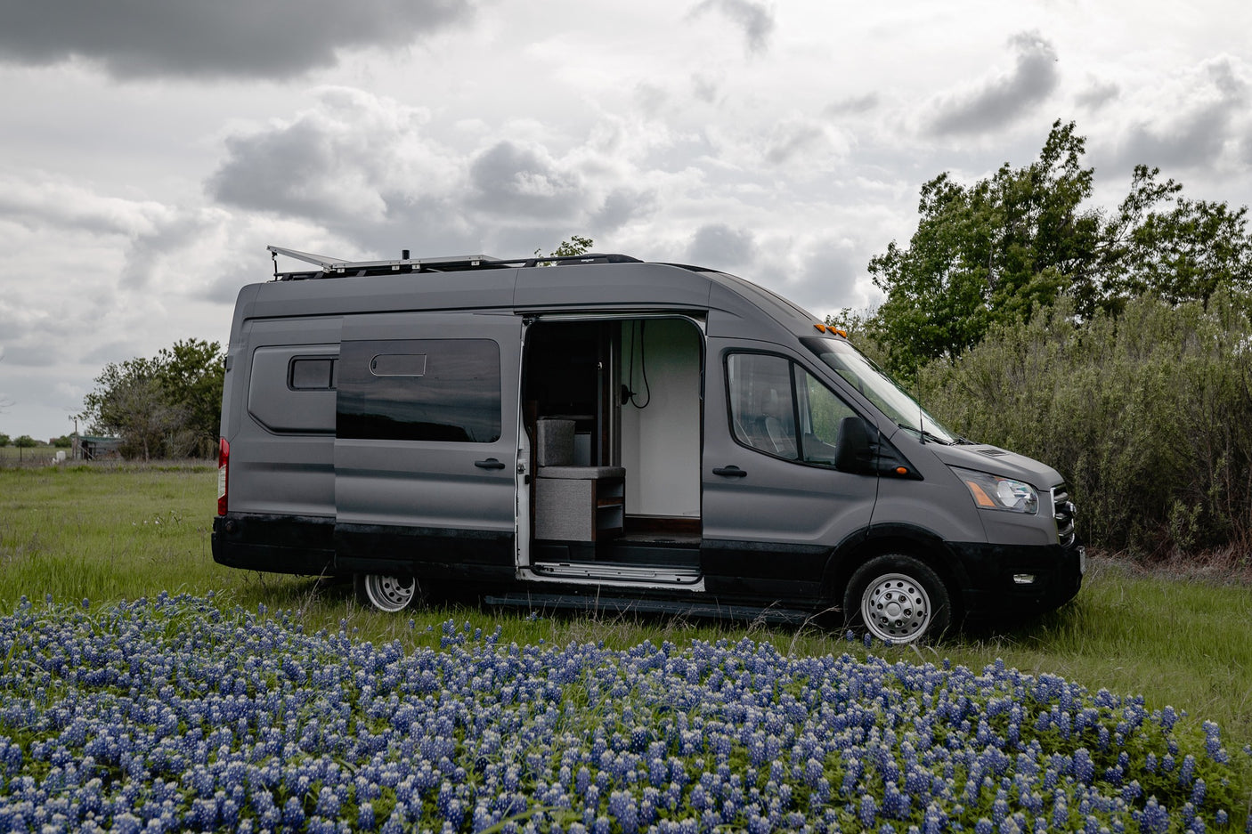 A grey camper van (RV rental) with its side door open is parked amidst a field of blue flowers under a cloudy sky.