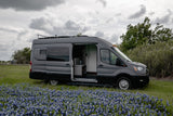 A grey camper van (RV rental) with its side door open is parked amidst a field of blue flowers under a cloudy sky.