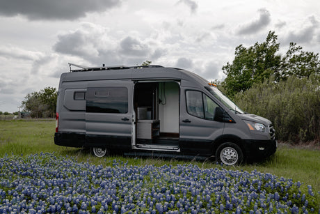 A grey camper van (RV rental) with its side door open is parked amidst a field of blue flowers under a cloudy sky.