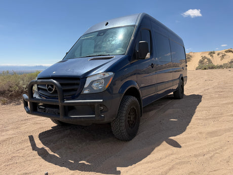 Dark blue Mercedes Sprinter van with bull bar and off-road tires on a sandy trail, perfect for an adventure RV rental.