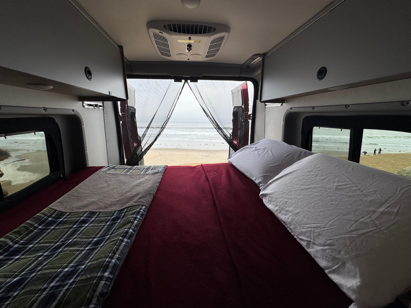 RV rental camper van interior. Features a cozy bed with blankets/pillows, and a clear beach/ocean view through open rear doors.