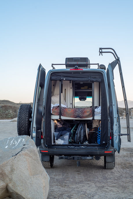 Open back of an RV rental camper van showing bed, gear storage, and rooftop AC unit. Equipped for adventure travel.