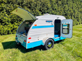 Compact Retro Jr. teardrop RV rental with open rear kitchen and door revealing cozy bed, on green grass.