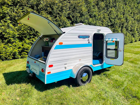 Compact Retro Jr. teardrop RV rental with open rear kitchen and door revealing cozy bed, on green grass.