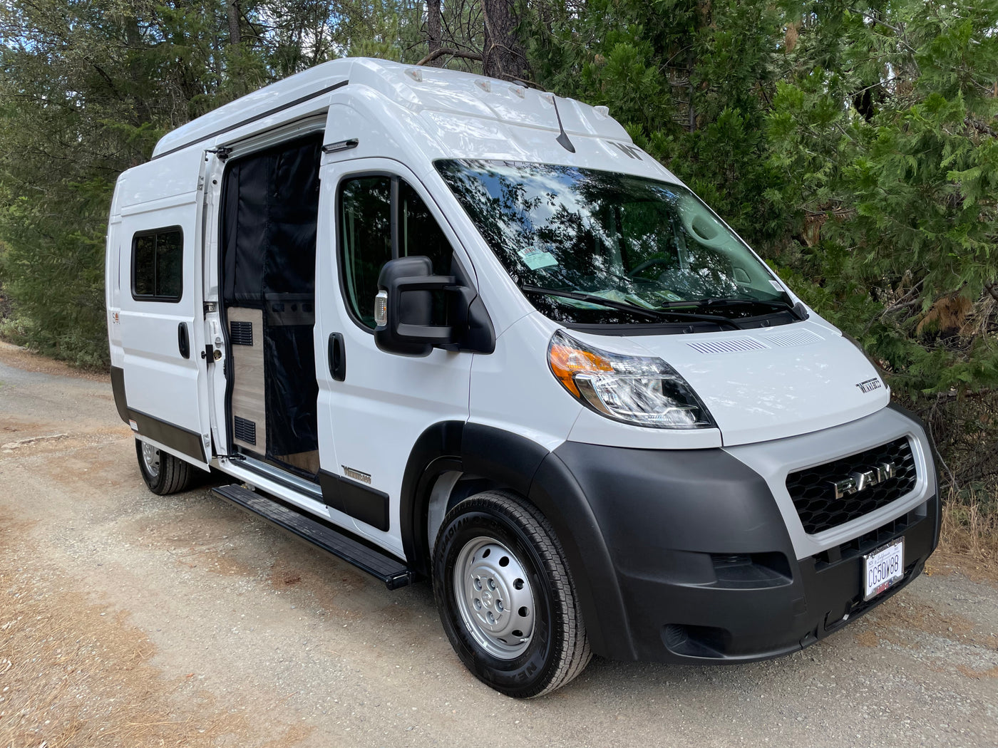 White Winnebago camper van rv rental parked on a dirt road, side door open. Perfect for forest adventures.