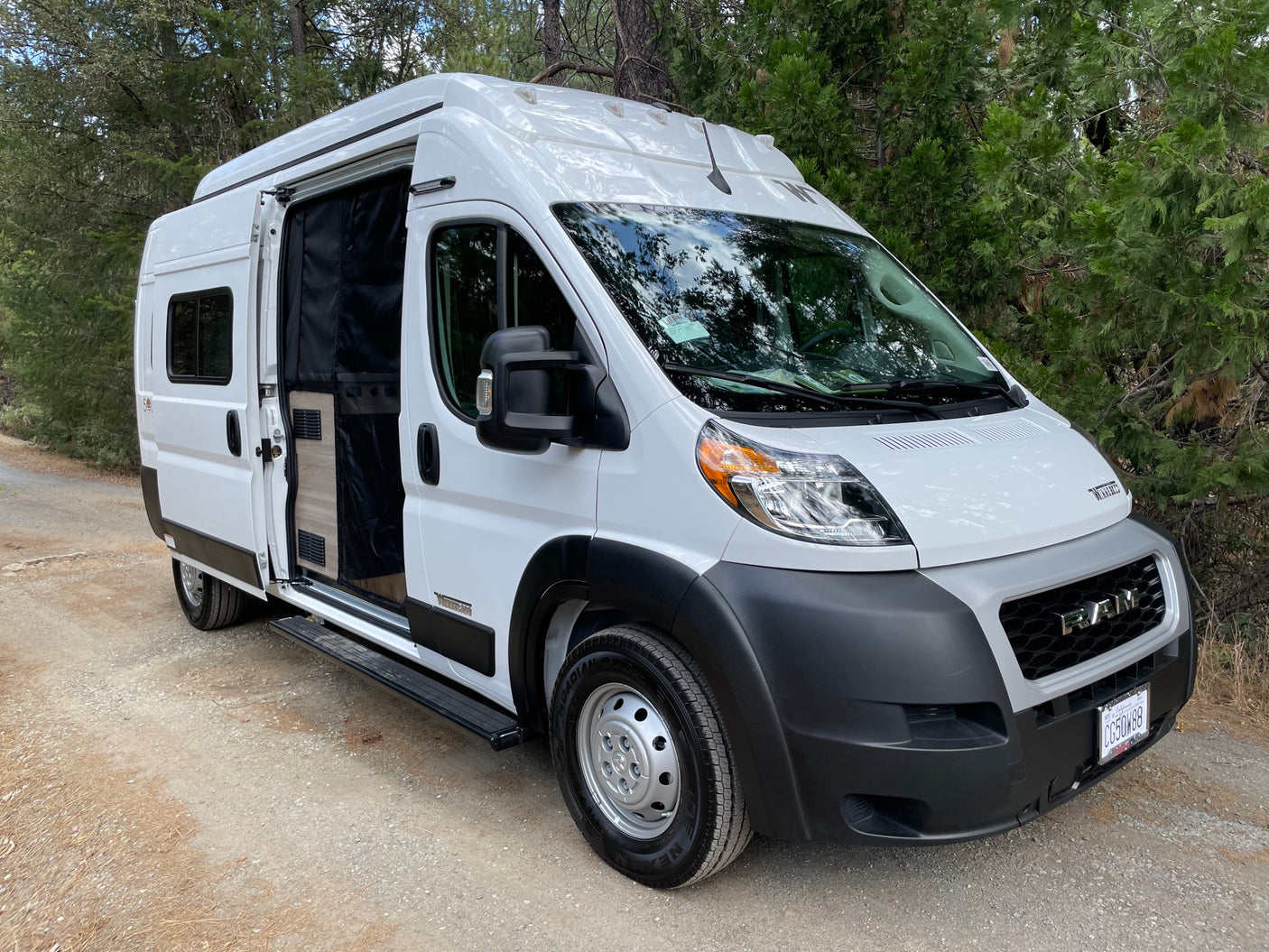 White Winnebago camper van rv rental parked on a dirt road, side door open. Perfect for forest adventures.