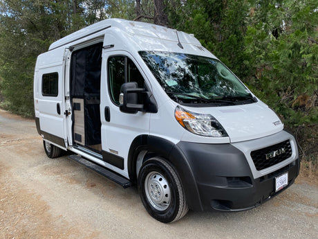 White Winnebago camper van rv rental parked on a dirt road, side door open. Perfect for forest adventures.