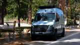 Light blue Winnebago Mercedes-Benz Sprinter RV rental camper van with roof rack, parked on a paved road in a pine forest.