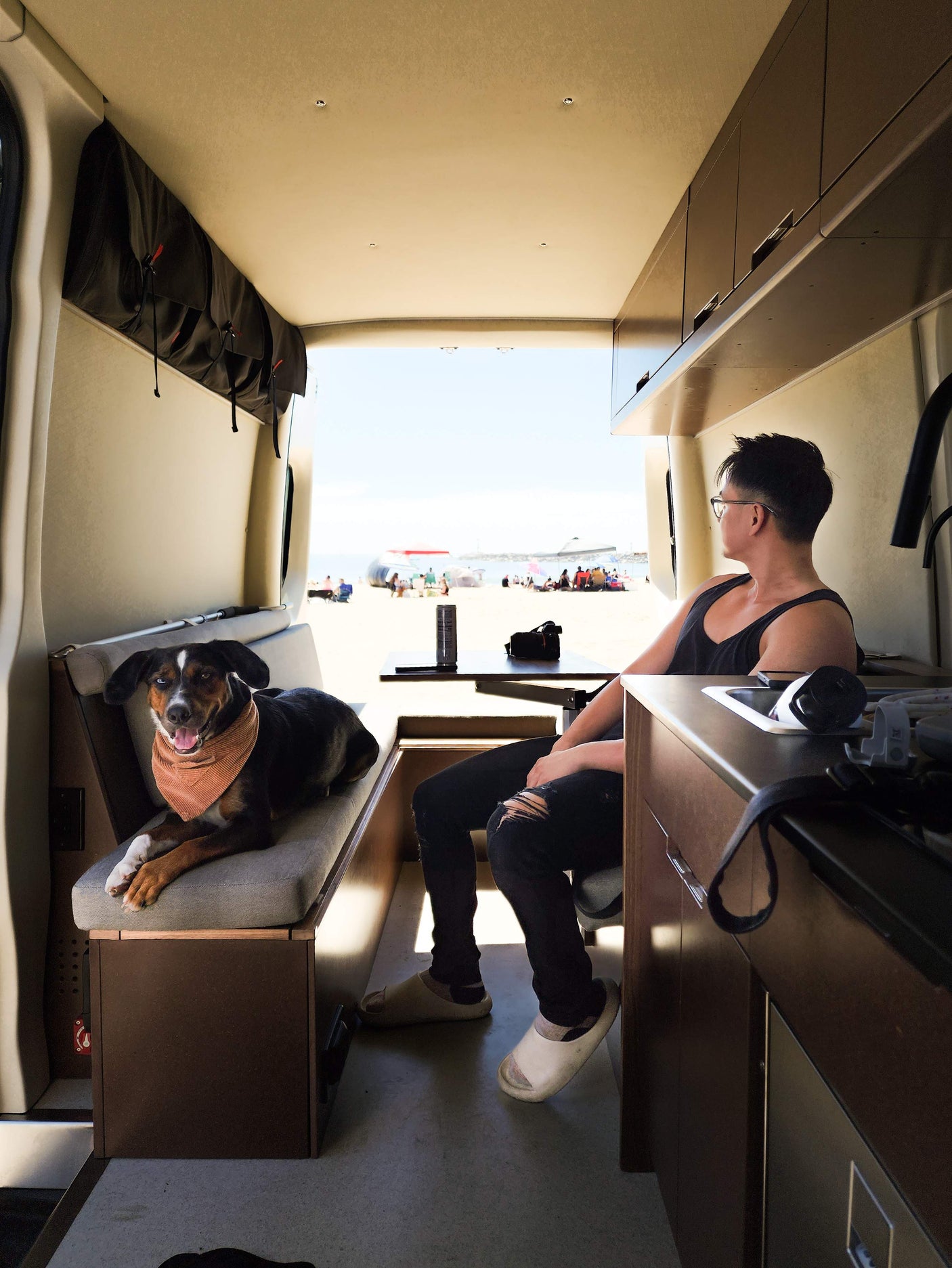Inside an RV rental: A man and dog enjoy beach views. Overhead storage & kitchen are visible, with people on the sand outside.