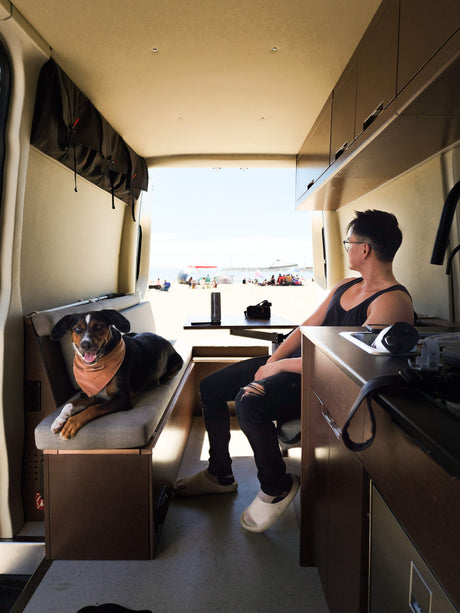 Inside an RV rental: A man and dog enjoy beach views. Overhead storage & kitchen are visible, with people on the sand outside.