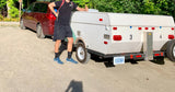 Man gives thumbs-up next to his compact RV rental trailer and minivan on a dirt road, ready for camping adventures.