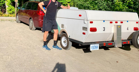 Man gives thumbs-up next to his compact RV rental trailer and minivan on a dirt road, ready for camping adventures.