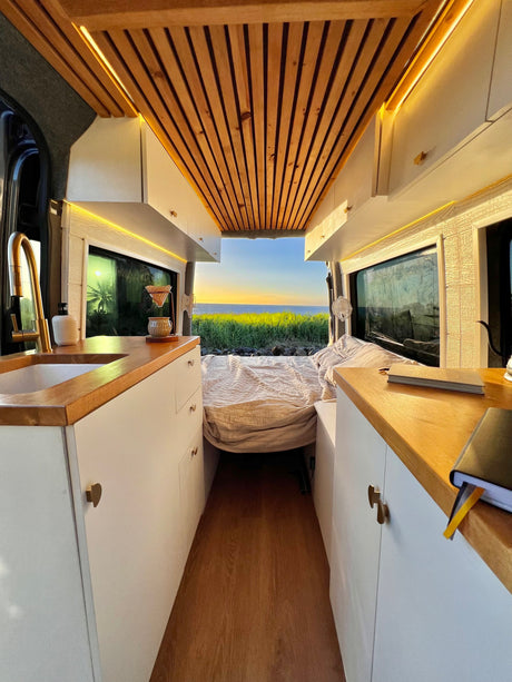 Cozy RV rental interior with wooden ceiling, functional kitchen & stunning ocean sunset view from the back. Perfect for van life adventures!