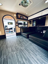 Tour this rv rental's interior: grey wood-look flooring, two dark sofa beds, a TV, and an open archway revealing the campsite.