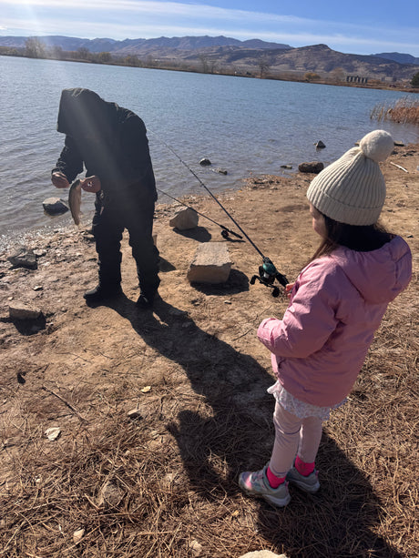 An adult holding a freshly caught fish, with a child and fishing rods by a scenic mountain lake. Perfect RV rental outdoor adventure.
