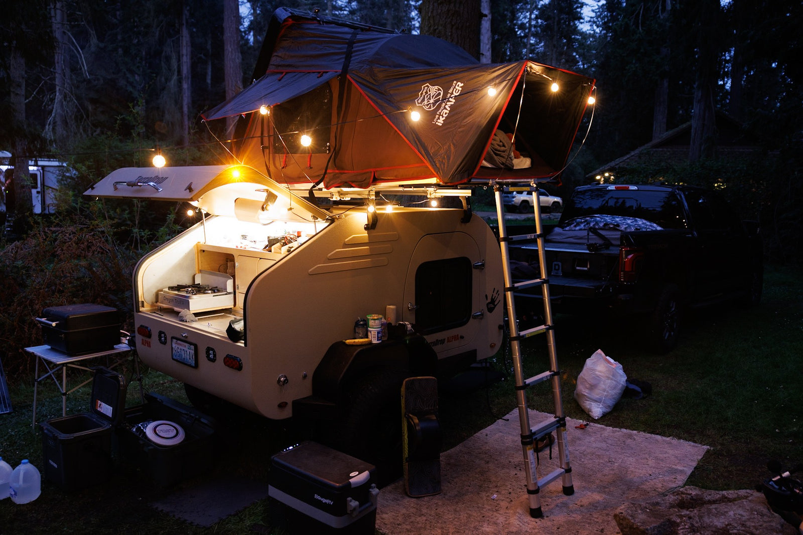  complete with a rooftop tent for off-road adventures.|Muddy motocross track winds through dense trees. An RV rental-style trailer setup and vans are parked trackside for the event.|Illuminated RV rental teardrop trailer with a cozy iKamper rooftop tent & outdoor kitchen
