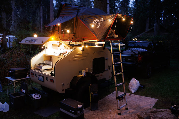  complete with a rooftop tent for off-road adventures.|Muddy motocross track winds through dense trees. An RV rental-style trailer setup and vans are parked trackside for the event.|Illuminated RV rental teardrop trailer with a cozy iKamper rooftop tent & outdoor kitchen