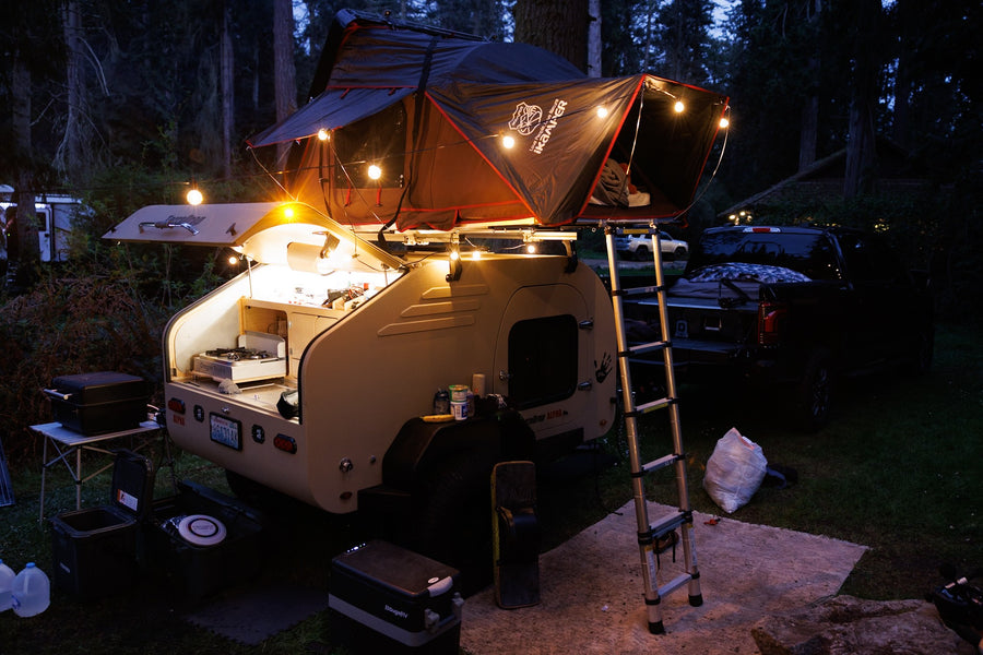  complete with a rooftop tent for off-road adventures.|Muddy motocross track winds through dense trees. An RV rental-style trailer setup and vans are parked trackside for the event.|Illuminated RV rental teardrop trailer with a cozy iKamper rooftop tent & outdoor kitchen