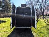 Rear view of a silver RV rental van with wide open doors revealing its interior and a bike rack, parked on green grass.