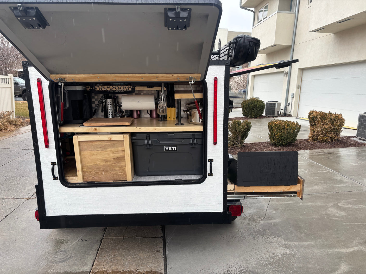 Compact RV rental: Rear view of an open camper trailer revealing a well-equipped outdoor camping kitchen with a cooler & pull-out counter.