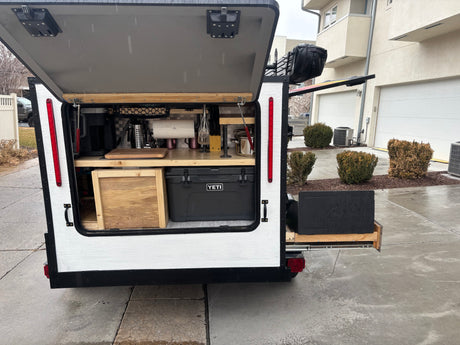 Compact RV rental: Rear view of an open camper trailer revealing a well-equipped outdoor camping kitchen with a cooler & pull-out counter.