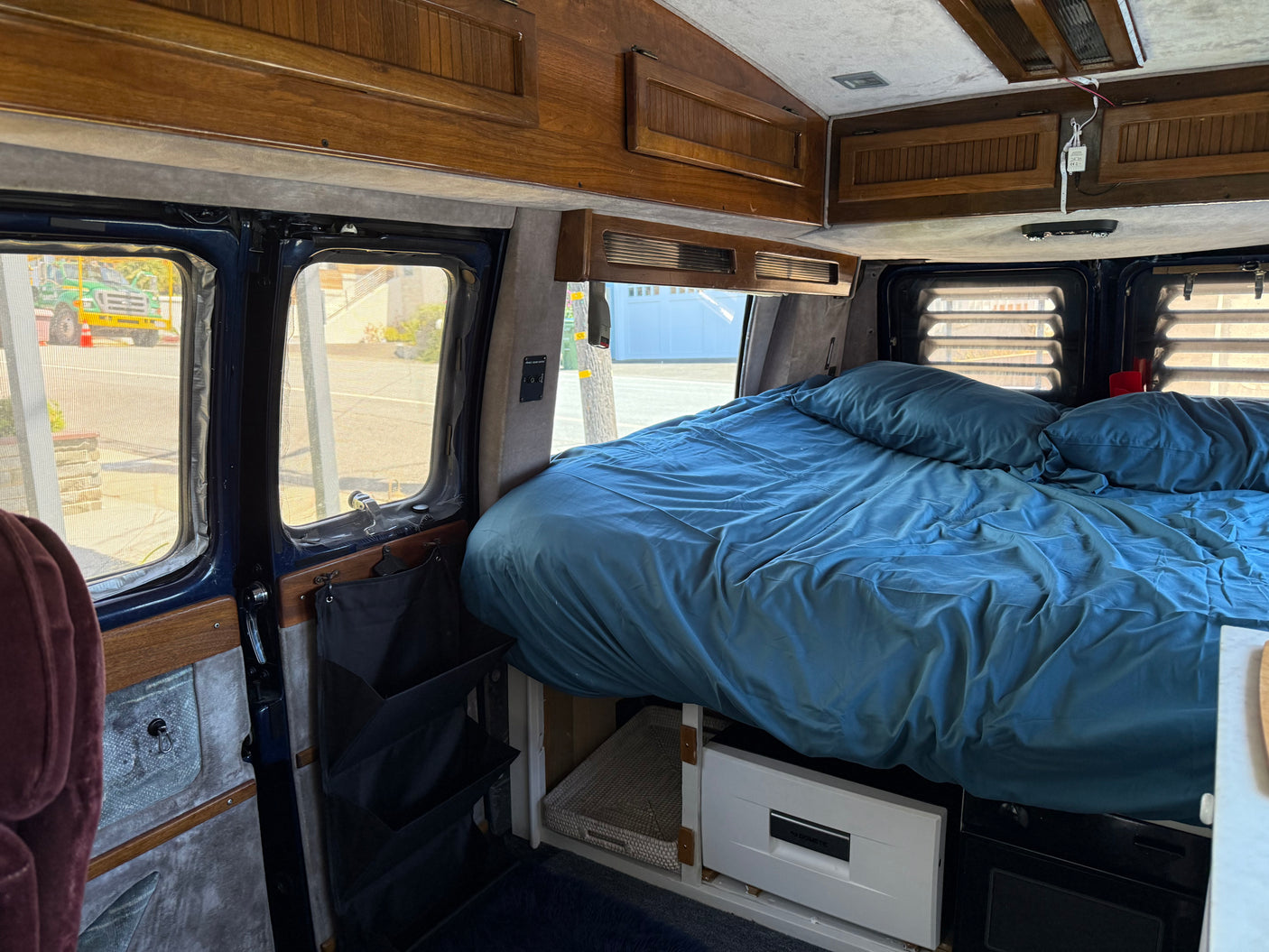 RV rental interior: comfy blue bed, wooden cabinets, window screens, and under-bed storage with a Dometic unit.