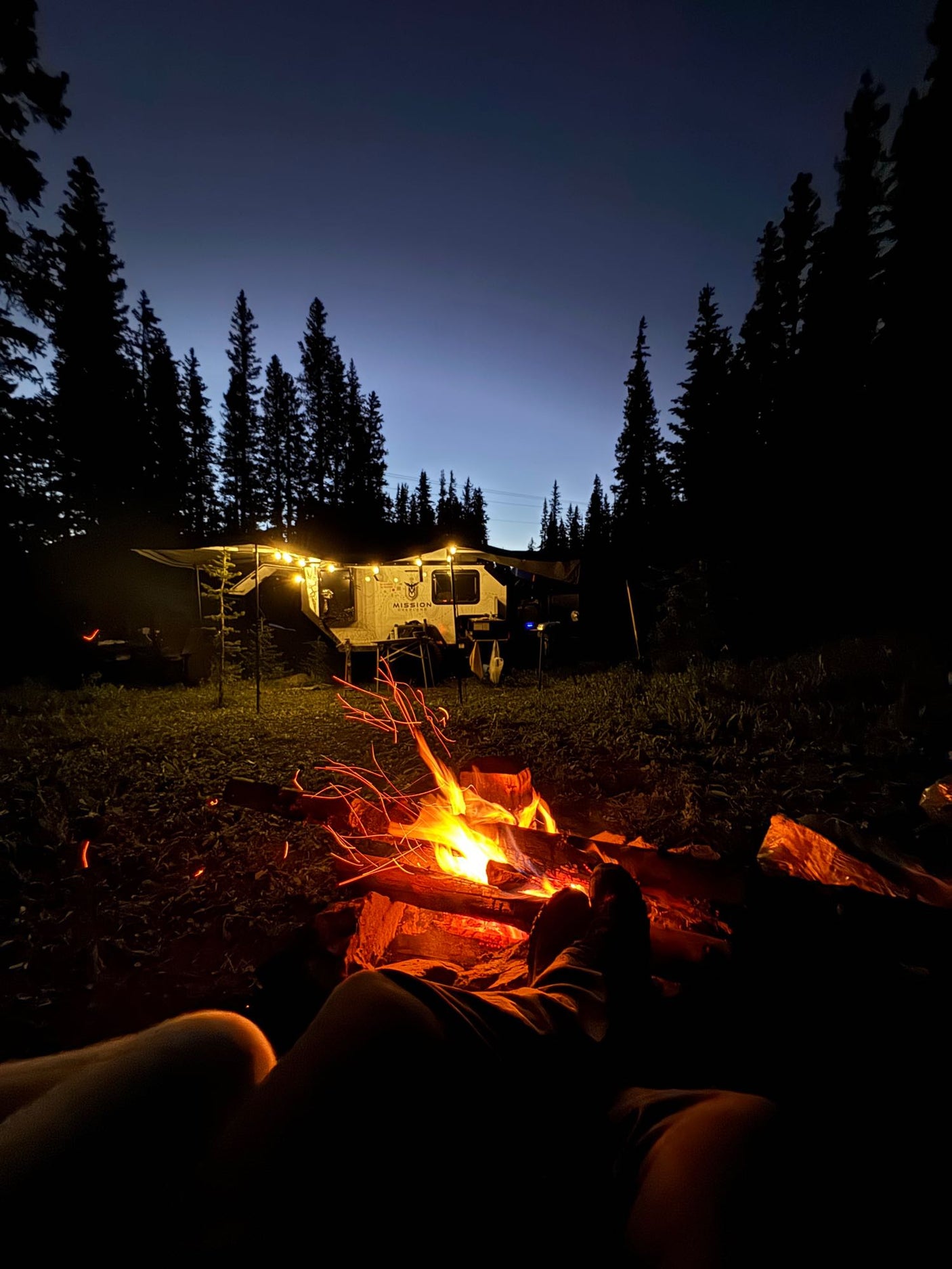 Cozy night camping: Relaxing by a campfire with an RV rental under string lights in a dark forest at dusk.