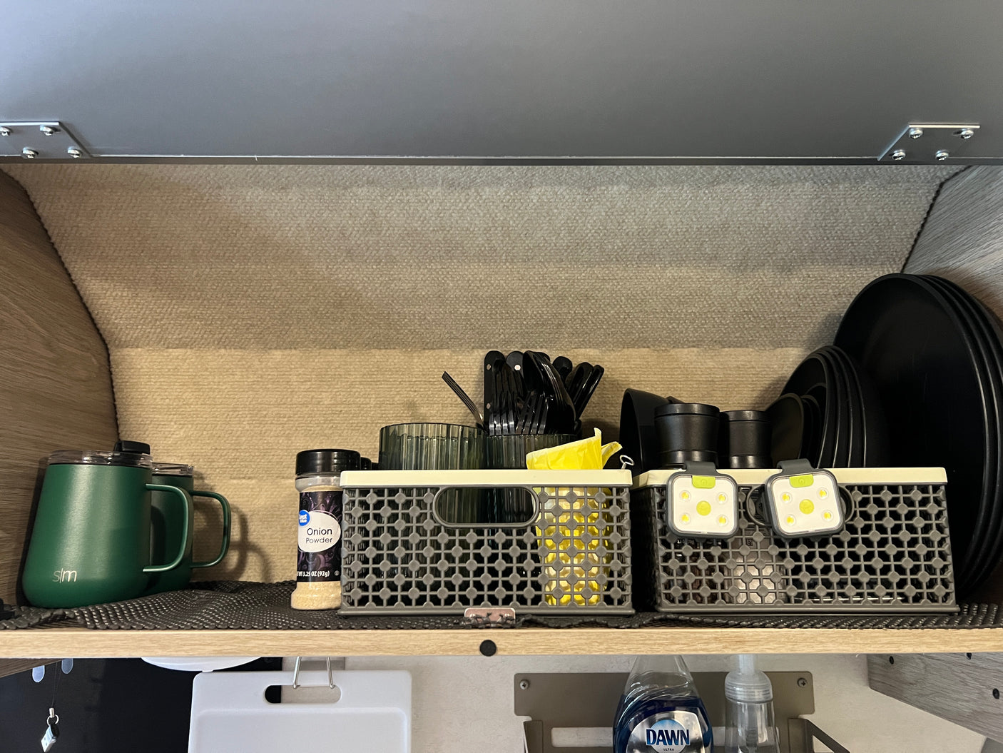 RV rental: An organized kitchen shelf holds green mugs, onion powder, baskets with utensils, cups, small lights, and black plates.