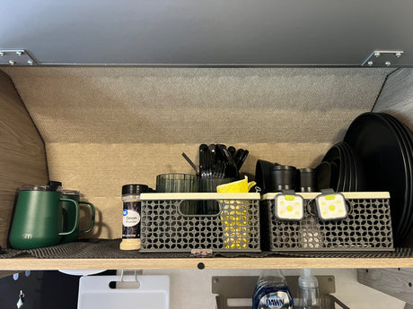 RV rental: An organized kitchen shelf holds green mugs, onion powder, baskets with utensils, cups, small lights, and black plates.
