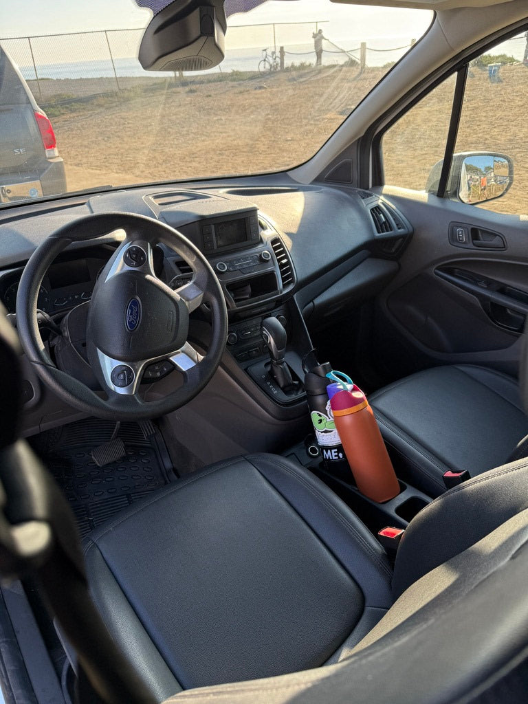 Inside an RV rental: The Ford van's dashboard, steering wheel, and two water bottles in the console, with a beach and ocean view outside.