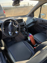 Inside an RV rental: The Ford van's dashboard, steering wheel, and two water bottles in the console, with a beach and ocean view outside.