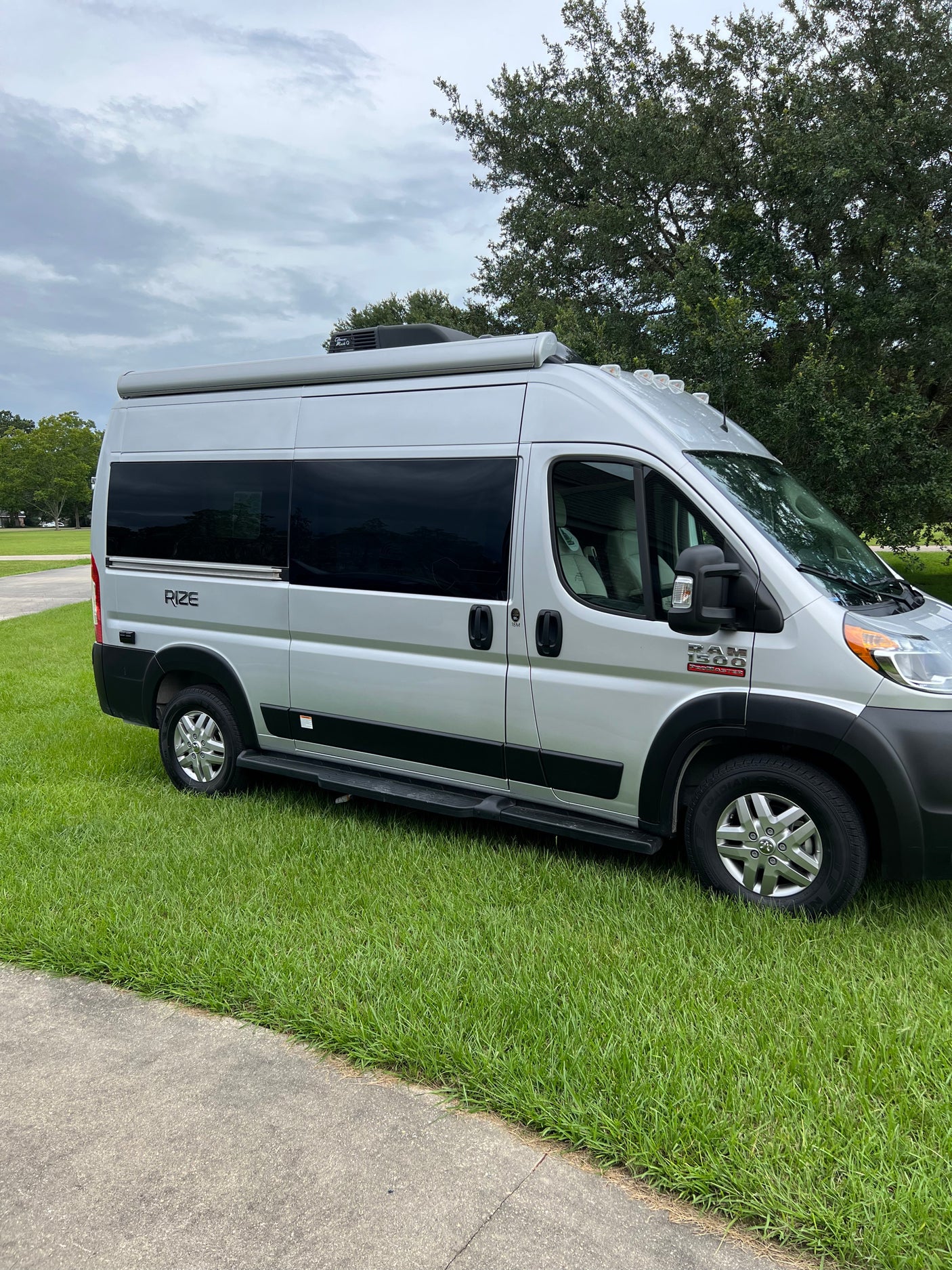 Silver Rize Ram ProMaster camper van RV rental, parked on grass beside a path under cloudy skies.