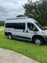 Silver Rize Ram ProMaster camper van RV rental, parked on grass beside a path under cloudy skies.