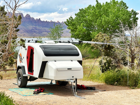 A sleek white RV rental teardrop trailer with an awning set up for camping near a lake with dramatic mountains.