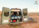  table/chairs outside.|Couple relaxing inside a camper van (RV rental) with its back doors open in a scenic desert landscape at dusk.|RV rental interior: A modern campervan with a bright dining area