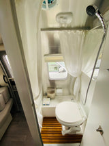Clean, compact RV rental bathroom showing a toilet, shower, wooden mat, and small window, ideal for comfortable travels.