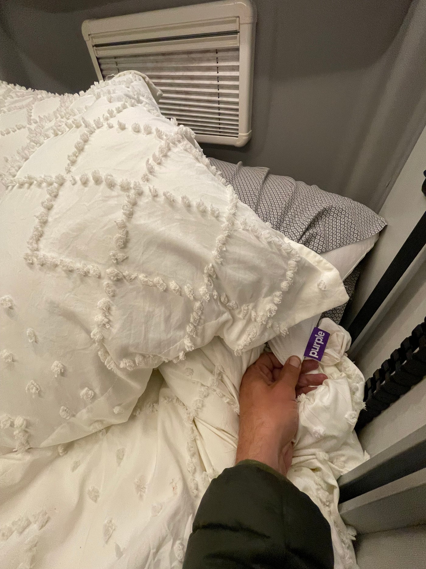 Hand holding Purple brand tag on textured white bedding in an RV rental. Window blinds & grey patterned pillowcase visible. — photo 6