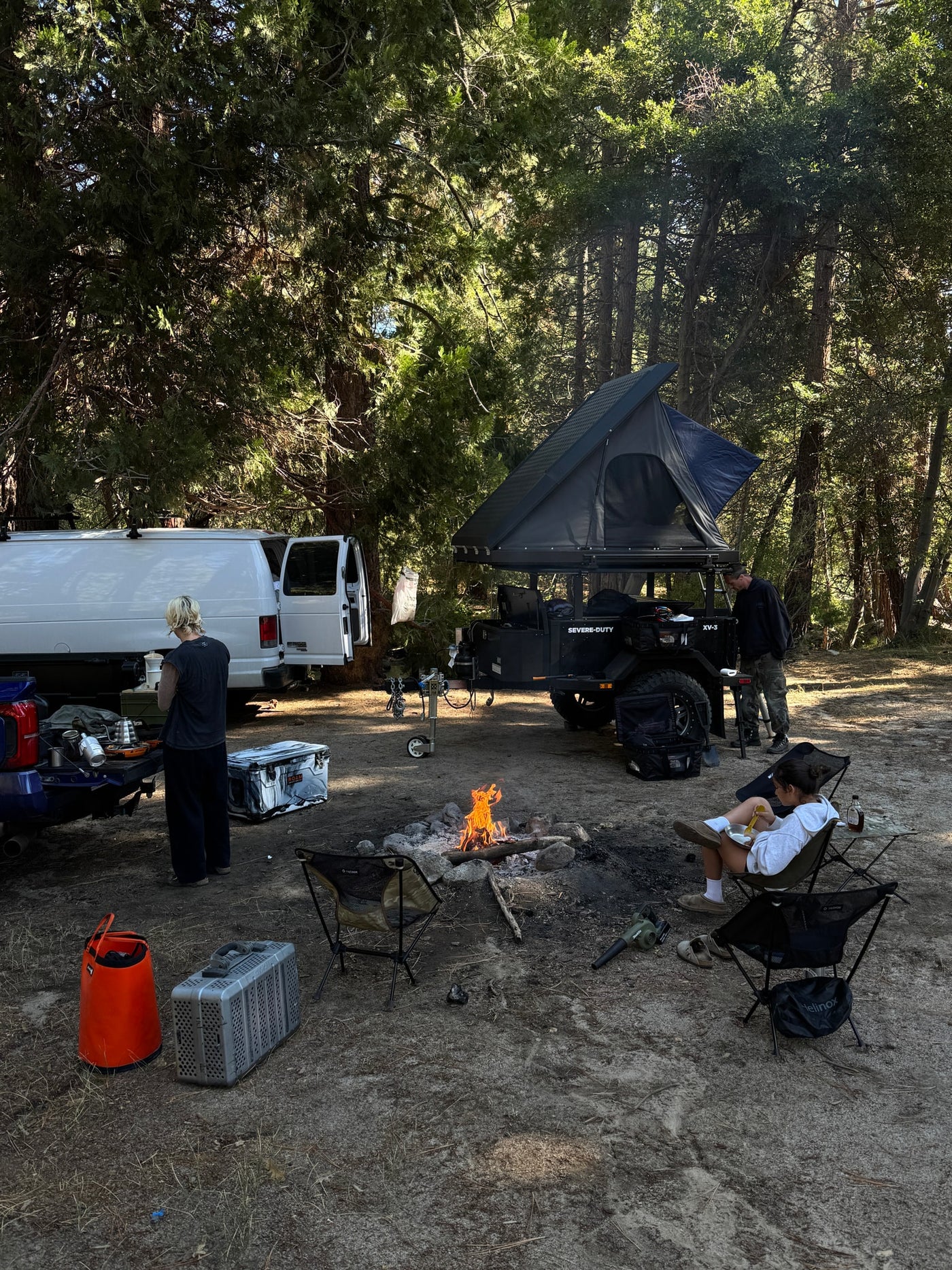 A serene campsite featuring an RV rental van, a Severe-Duty overland trailer with rooftop tent, and campers enjoying a warm campfire. — photo 4
