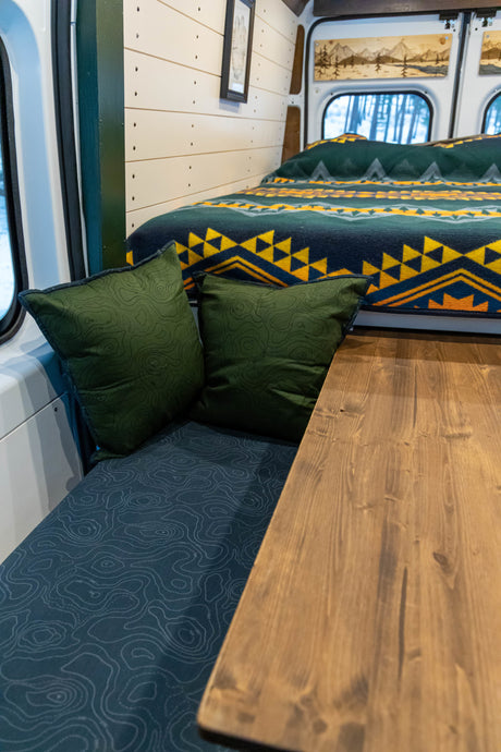 Experience an RV rental! Inside, find a wooden table, cozy bed with patterned blanket, and seating with green topo-map cushions.